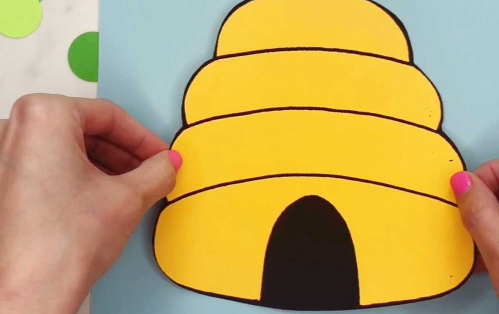 Hands hold a large yellow paper beehive with black outlines and a dark entrance opening. The beehive is positioned on a blue background.