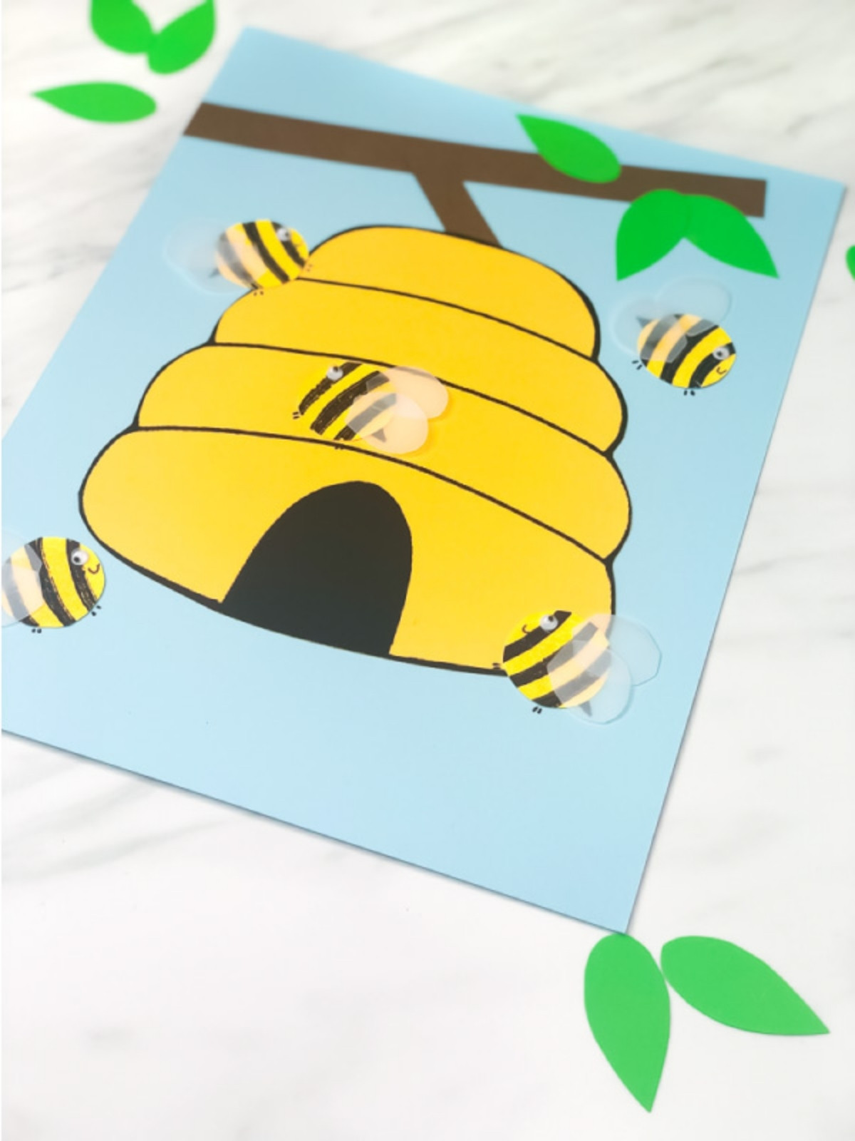 Finished paper craft of a yellow beehive on a blue background with several striped bees flying around it. Green paper leaves and a brown branch are placed above.