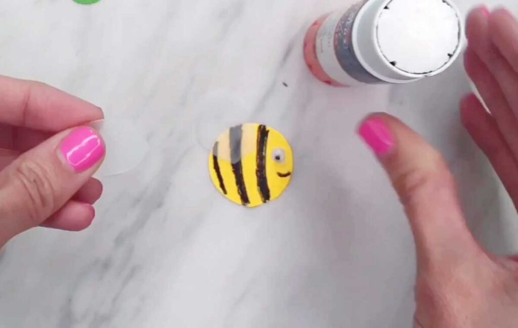 A small yellow paper circle with black stripes and a drawn smiley face is shown near a bottle of glue. The piece resembles a simple bee body.