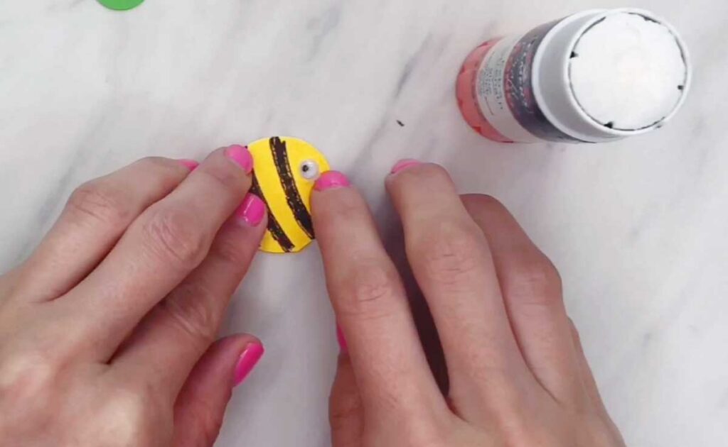Hands press a googly eye onto the striped yellow circle to complete the bee face. The bee’s features become more defined.