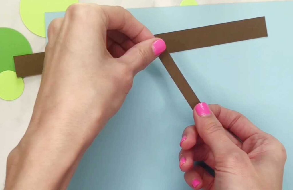 Hands attach brown paper strips diagonally across a blue background. The strips resemble branches or stems.