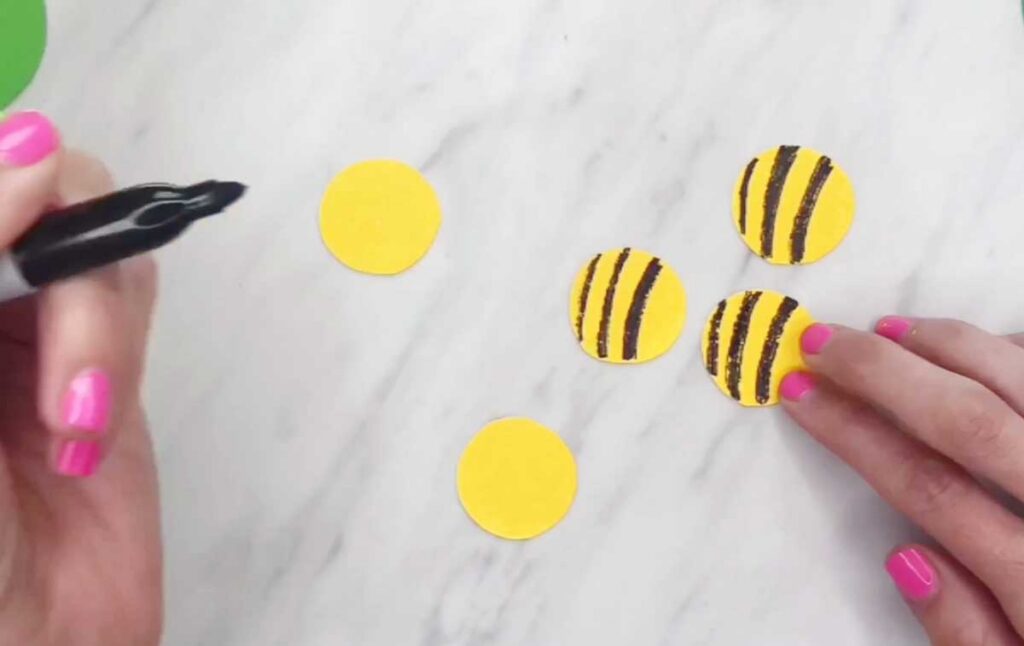 Several yellow paper circles are decorated with black stripes using a marker. The circles are being turned into multiple bee bodies.