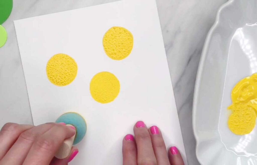 Hand uses a round sponge to stamp yellow paint circles onto white paper. Three textured yellow circles are visible on the page.
