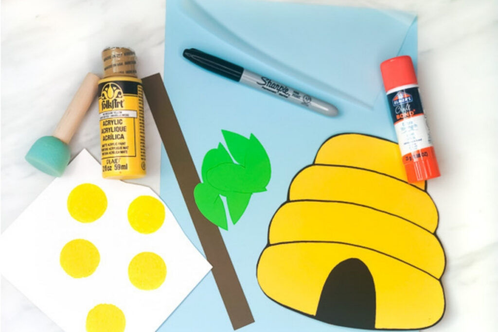 Craft materials arranged on a table including yellow paint, sponge stamp, paper cutouts, marker, glue stick, and a finished beehive template.