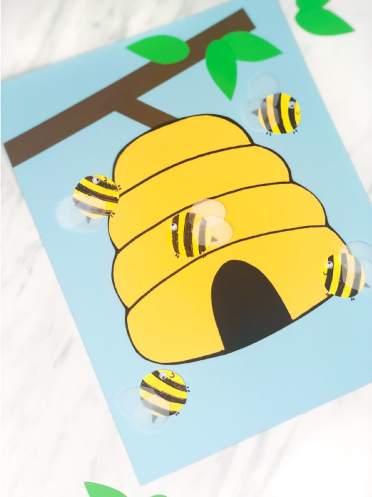 Close-up angled view of the paper beehive craft with multiple small bees attached around it. A brown paper branch and green leaves appear at the top.
