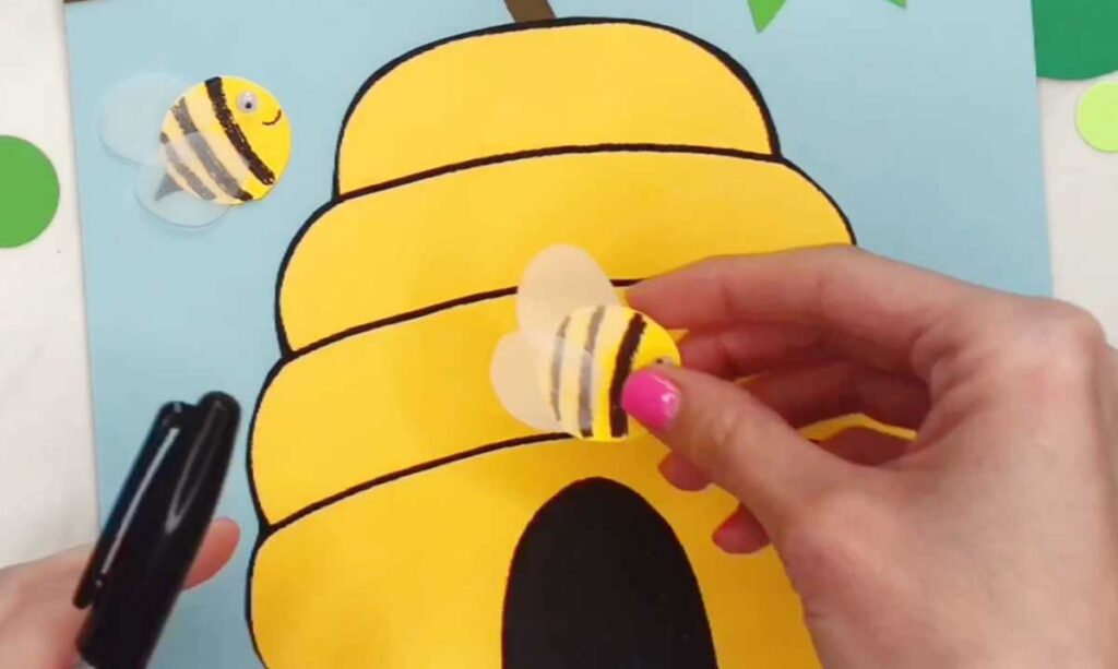 A finished paper bee with wings is held in front of a large yellow beehive on a blue background. Another bee is already attached near the hive.