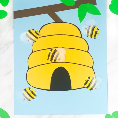 Paper craft of a yellow beehive hanging from a brown branch with green leaves on a blue background. Cutout bees with striped bodies and translucent wings are arranged around the hive.