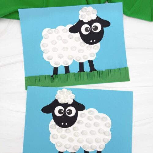 Two identical sheep crafts featuring a fluffy pom-pom body and black paper details rest on a table with a green fabric backdrop.