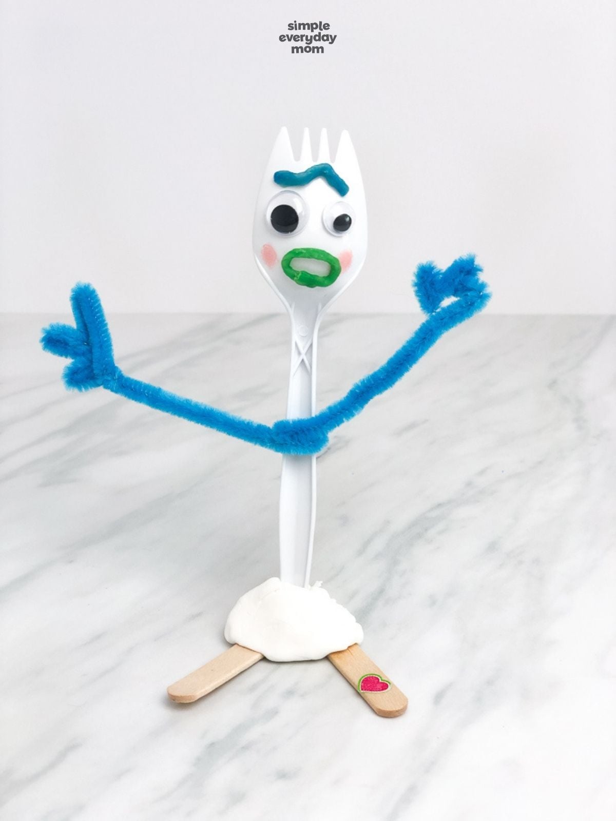 Finished Forky craft with blue pipe-cleaner arms, googly eyes, a green mouth, clay base, and craft-stick feet.