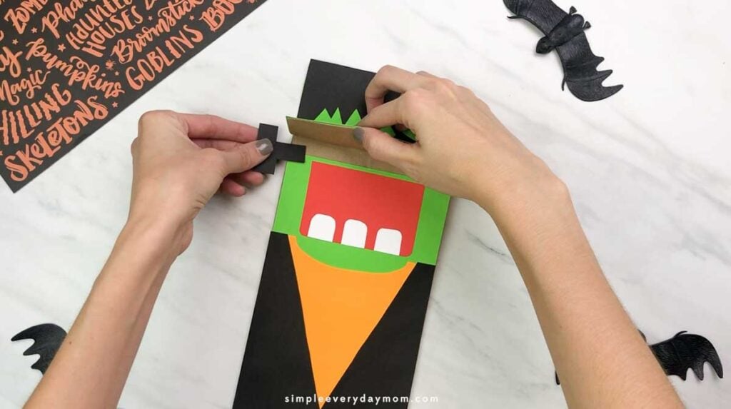 Hands attach a black paper neck bolt to the side of the Frankenstein paper bag puppet.