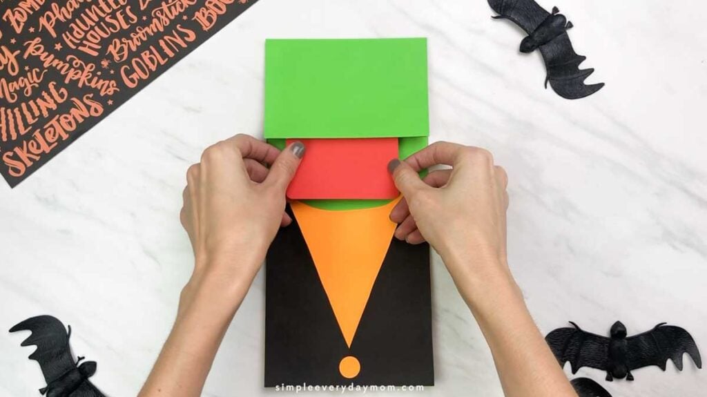 Hands position a red paper mouth beneath the green flap of the puppet’s face.