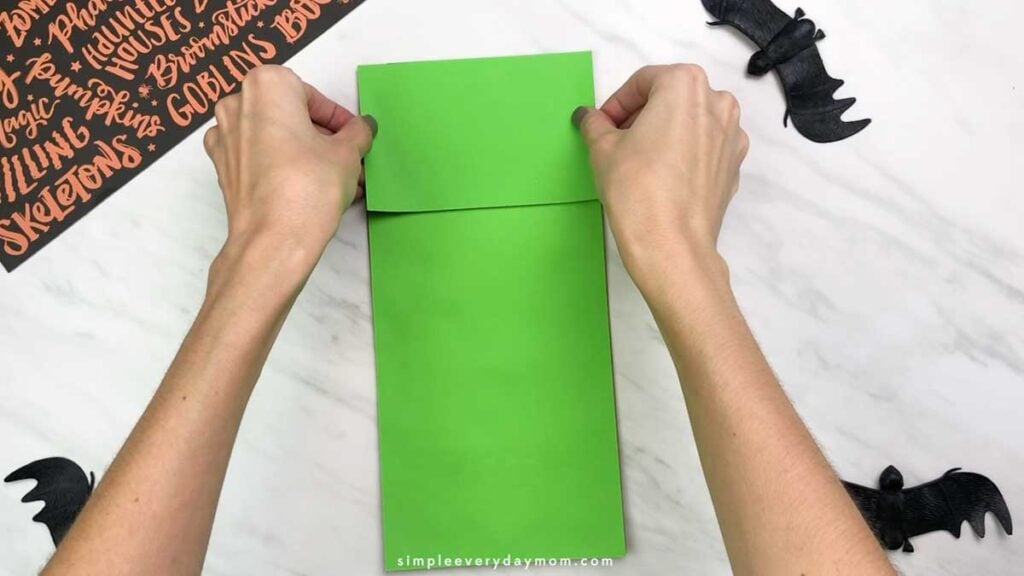 Hands fold the top flap of green paper over the paper bag to form the puppet’s head.