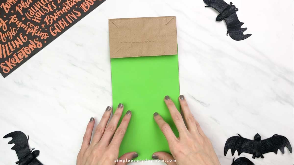 Hands position green paper over a brown paper bag to begin a Frankenstein puppet craft.