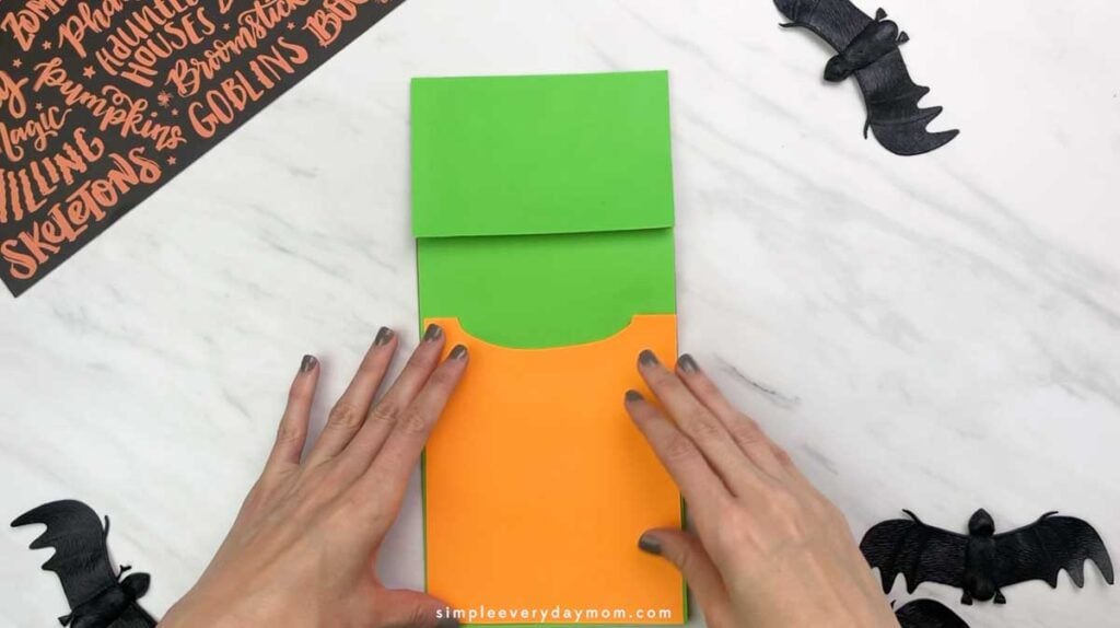 Hands place an orange paper shirt piece onto the green-covered paper bag.