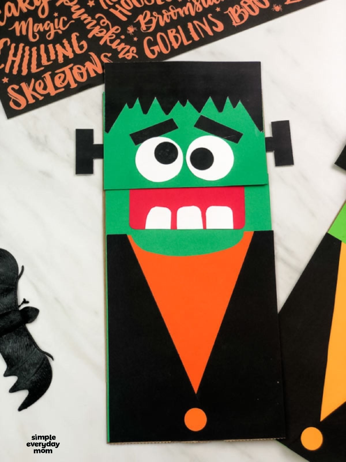 Front view of a handmade Frankenstein paper bag puppet with green skin, black hair and jacket, and an orange shirt.