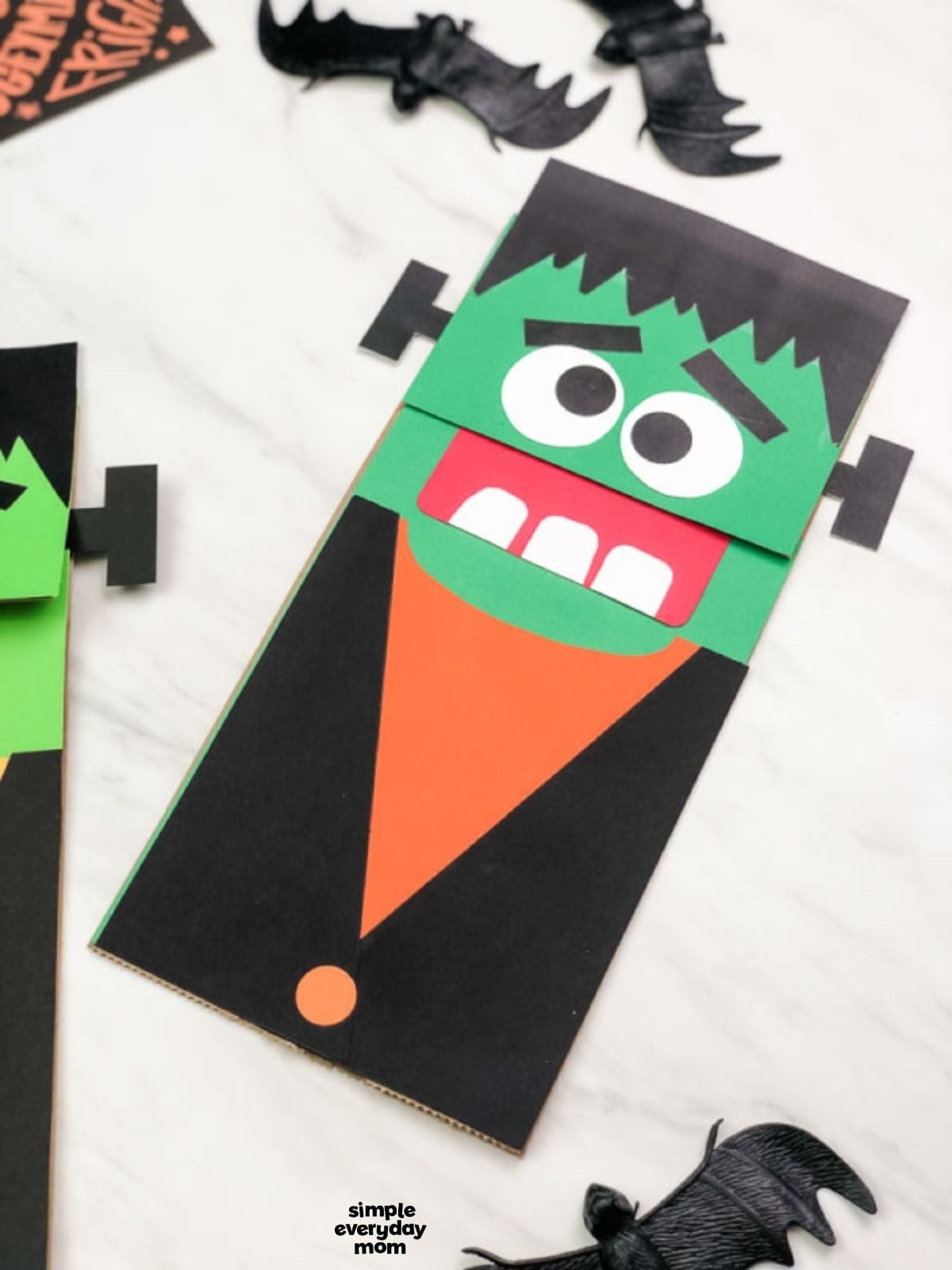 Close-up of a Frankenstein paper bag puppet with a green face, black hair, large eyes, red mouth, and orange shirt.
