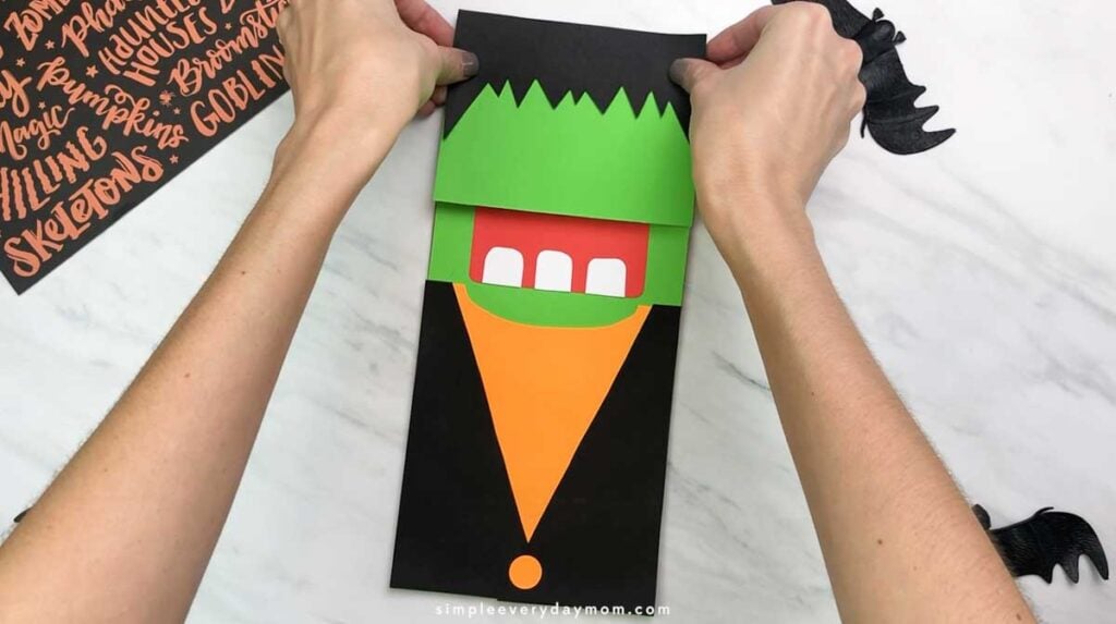 Hands place a black jagged hair piece along the top of the green Frankenstein face.
