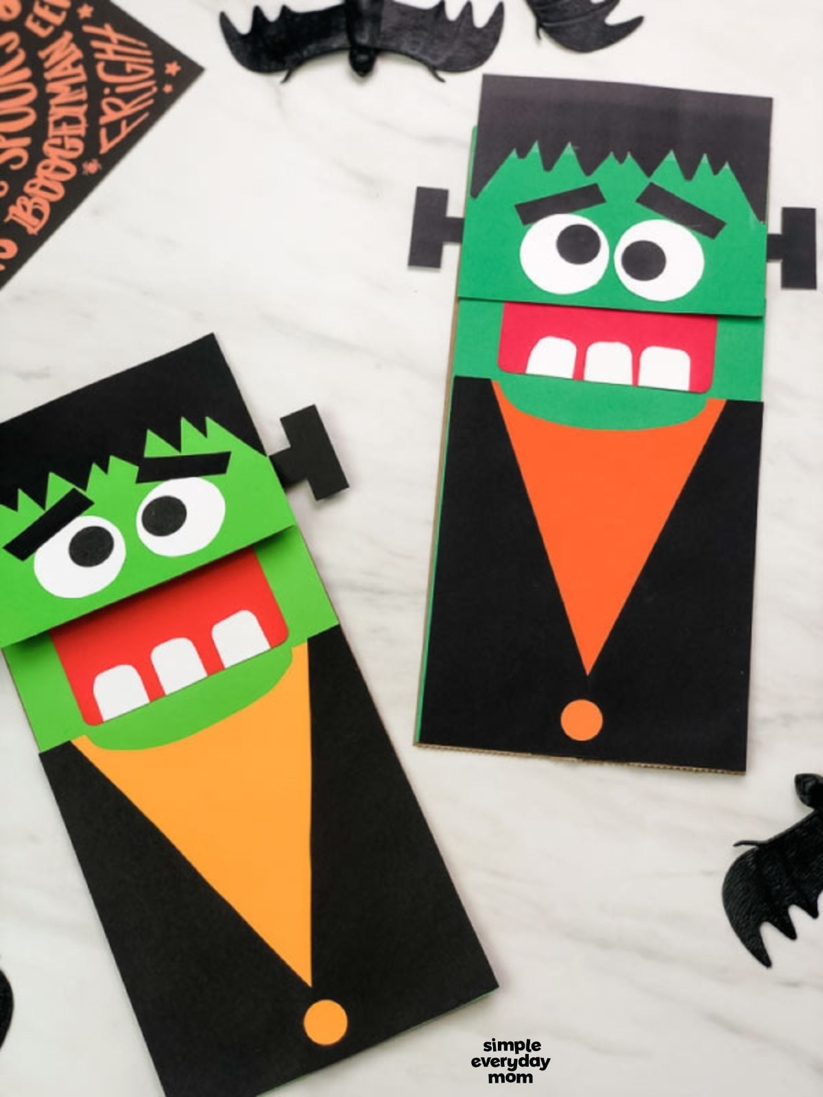 Two handmade Frankenstein paper bag puppets with green faces, black hair and jackets, orange shirts, and large white eyes.