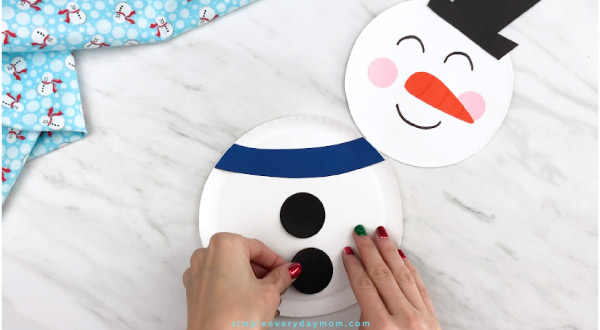 Hands gluing coal piece onto paper plate snowman 