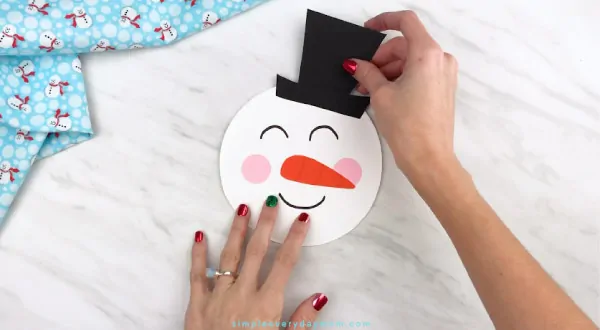Hands gluing hat onto paper plate snowman