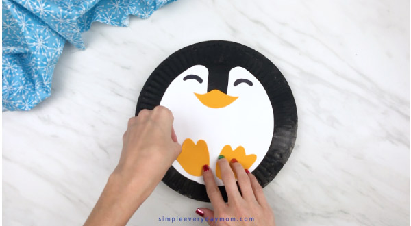 Hands gluing penguin feet to paper plate 
