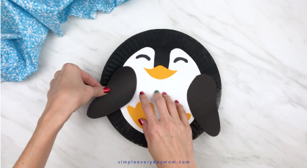 Hands gluing penguin wings to paper plate 