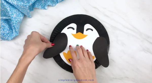 Hands gluing penguin wings to paper plate 