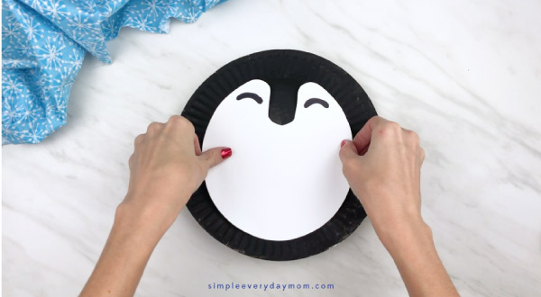 Hands gluing penguin body to paper plate 