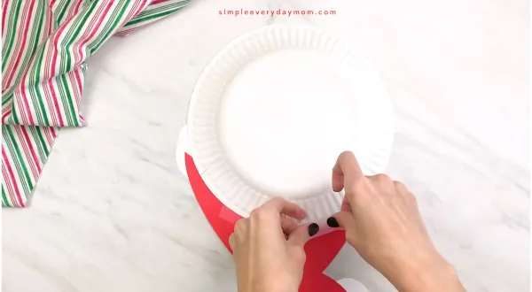 hands taping paper Santa hat onto Santa paper plate craft