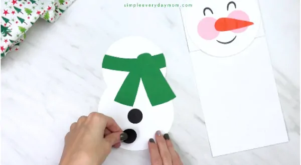 Hands gluing black circles onto paper bag snowman body 