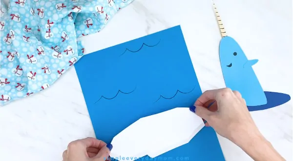 Hands gluing paper iceberg to blue paper 