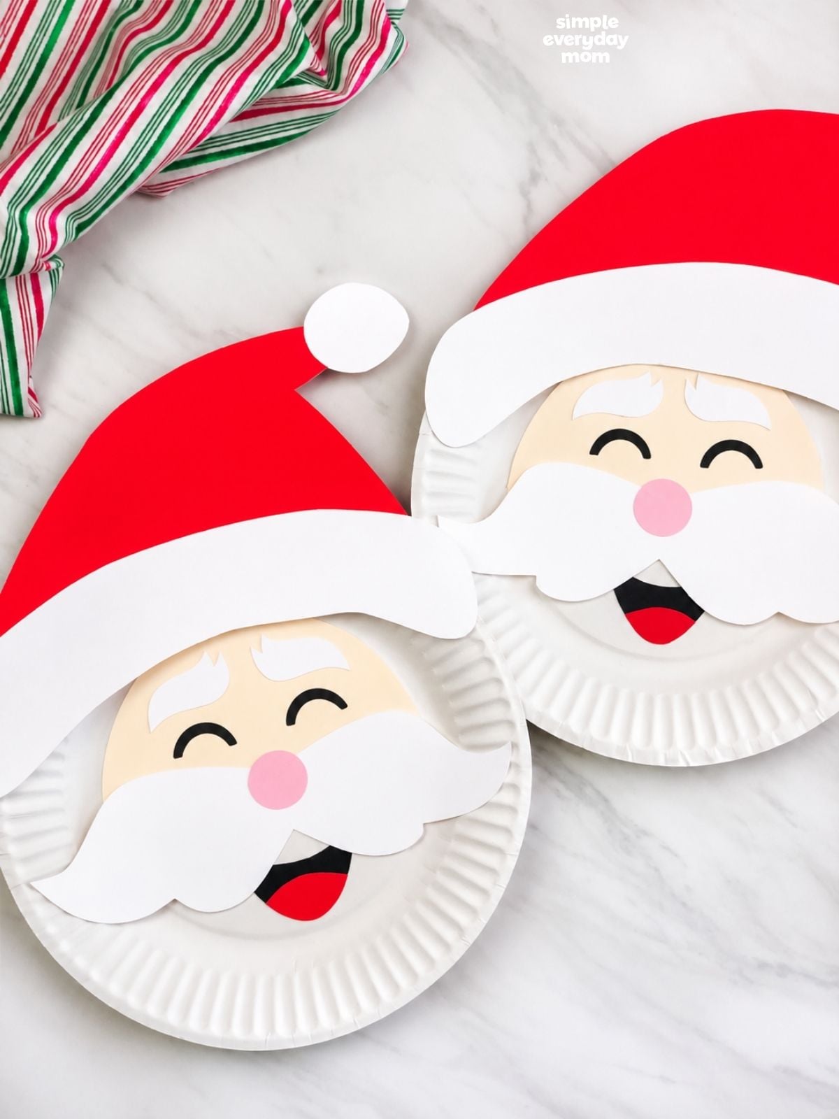 Two Santa Claus paper plate crafts with red hats, white mustaches, and smiling faces overlap on a white marble surface beside red, green, and white striped fabric.