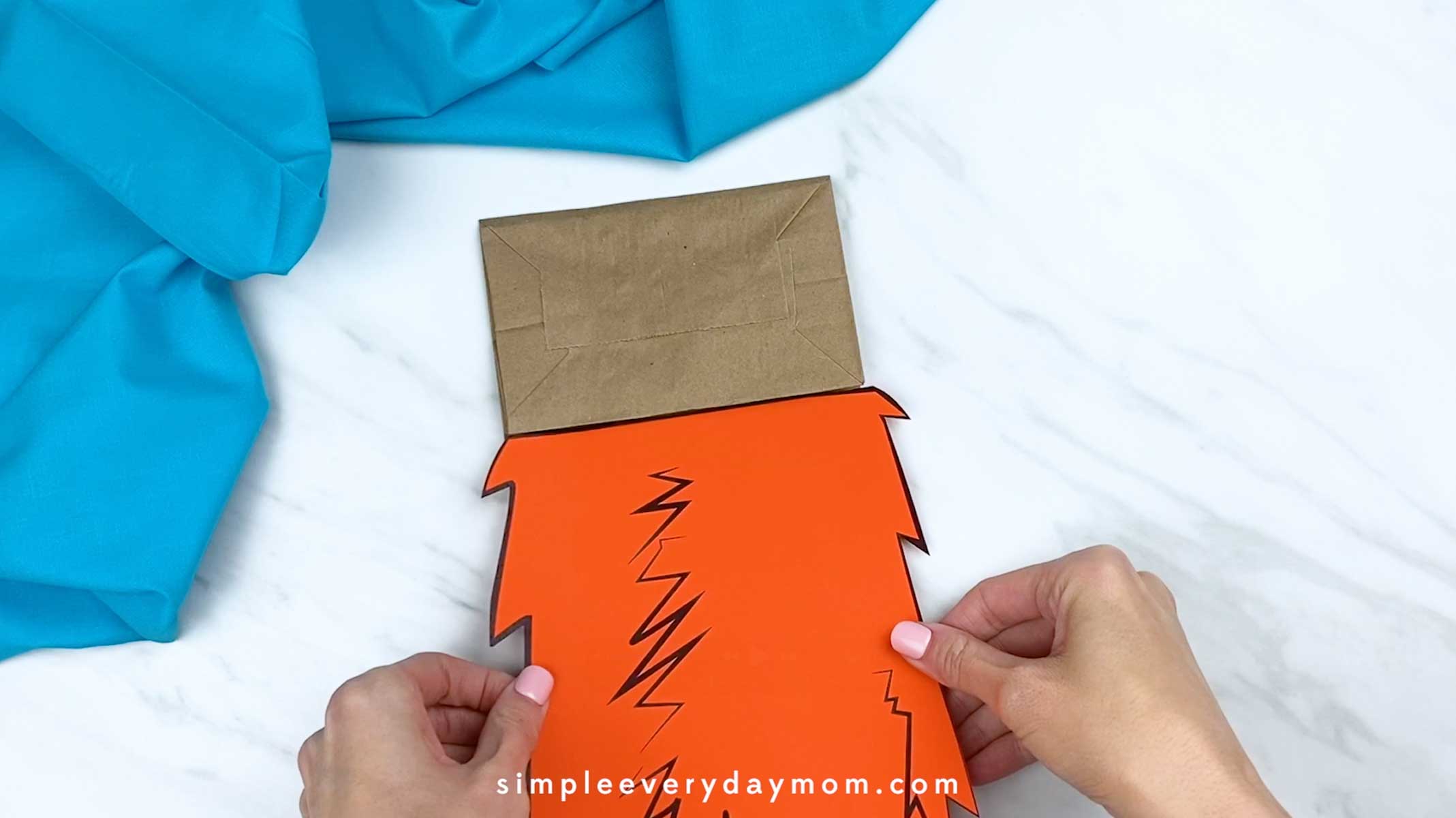 Hands placing an orange paper body piece onto a brown paper bag to begin assembling a fox craft.