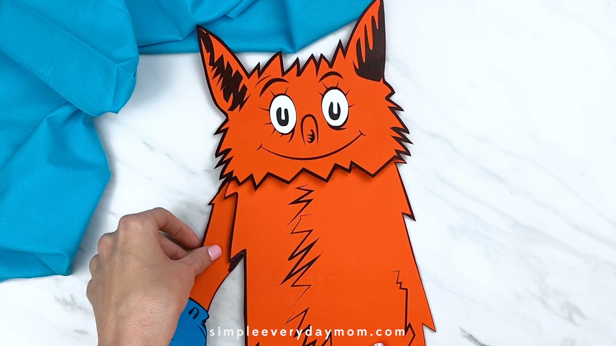 Hands attaching an orange arm piece with a blue glove to the side of the fox’s body.