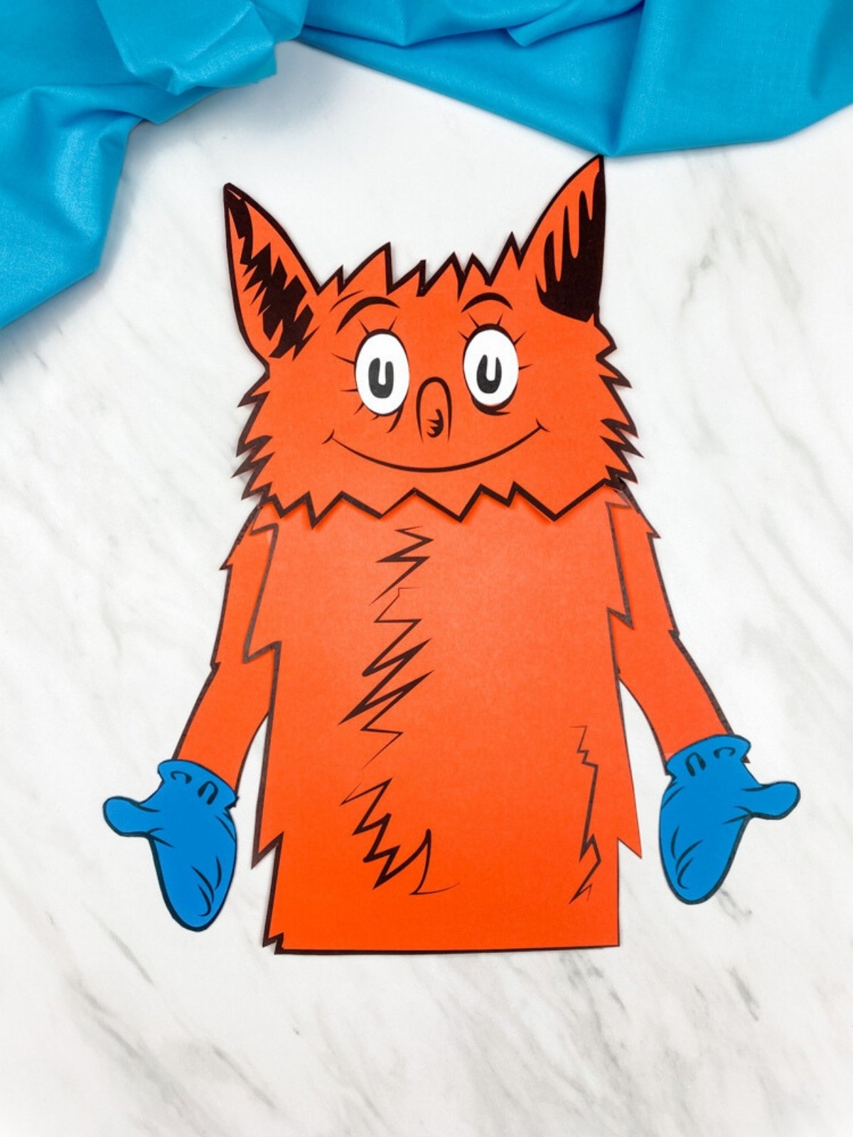 Single orange fox paper bag puppet centered on a white marble background with blue fabric at the top edge.