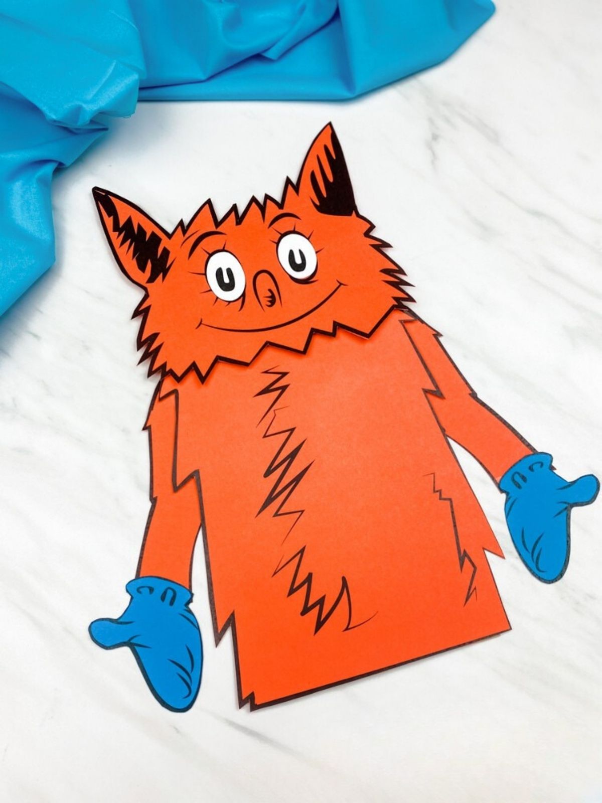 Finished orange fox paper bag puppet with blue mittens displayed on a white marble background with blue fabric in the corner.