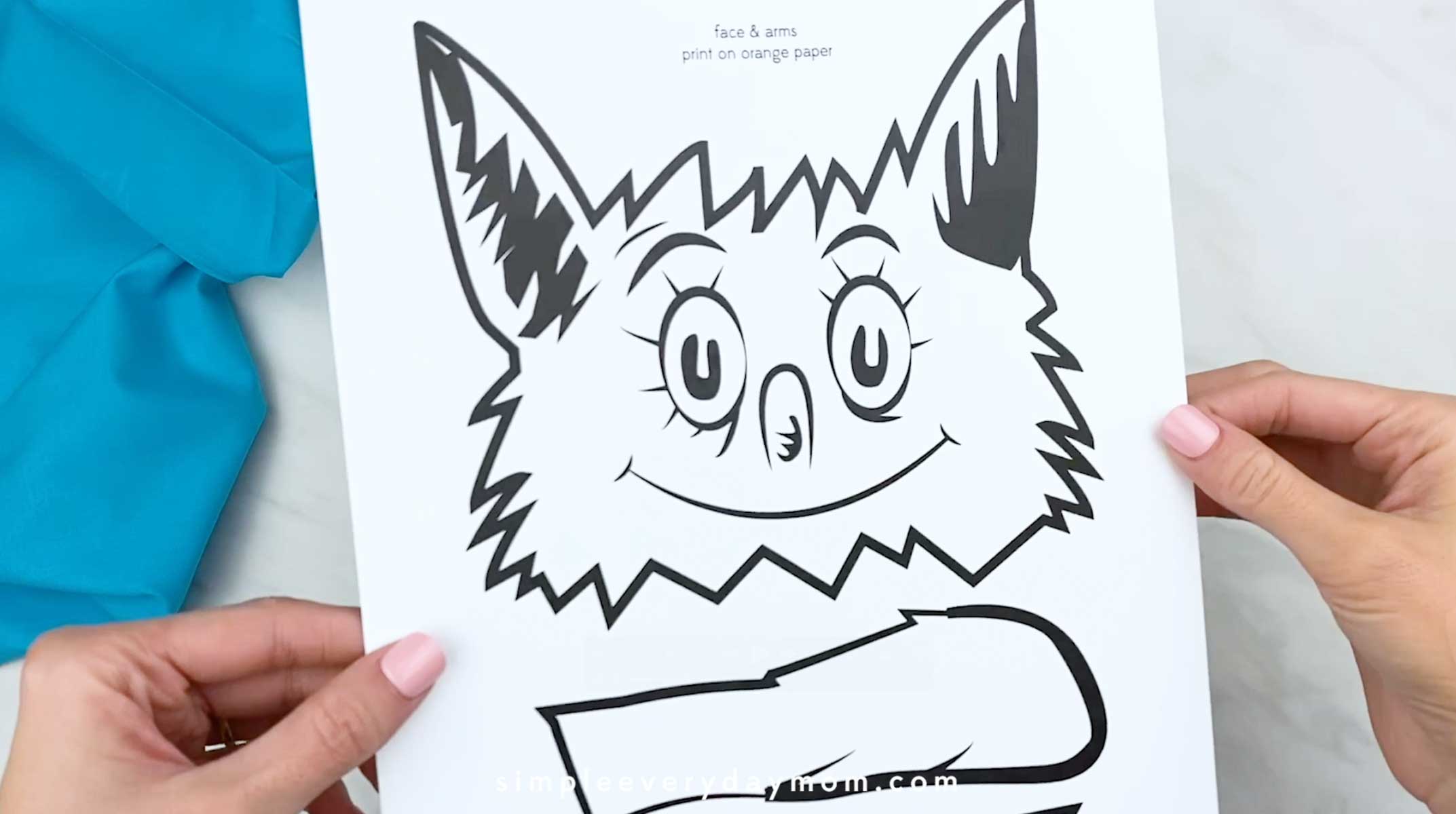 Hands holding a printed fox face and arms template on white paper over a light surface with blue fabric in the background.