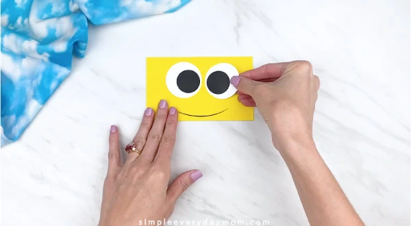 Hands gluing eyes onto yellow rectangle 