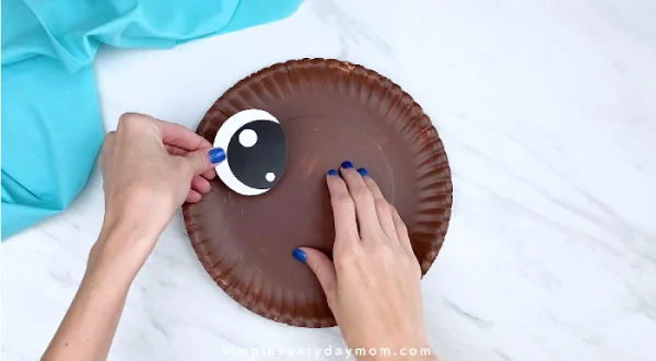 Hand gluing eye to brown paper plate 