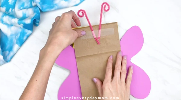 Hands taping pipe cleaner antennae to back of paper bag butterfly 