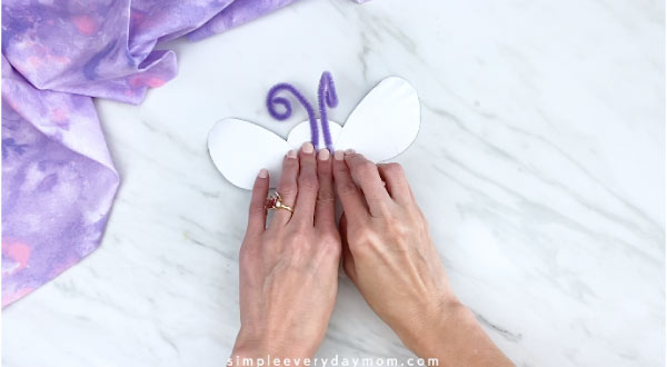 Hands tapping purple pipe cleaners to back of paper plate butterfly 