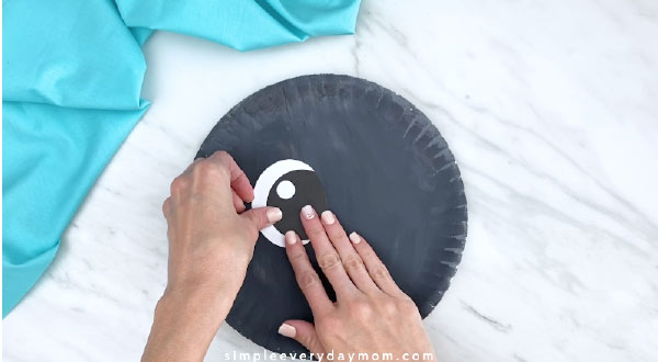 Hand gluing eye onto gray paper plate 