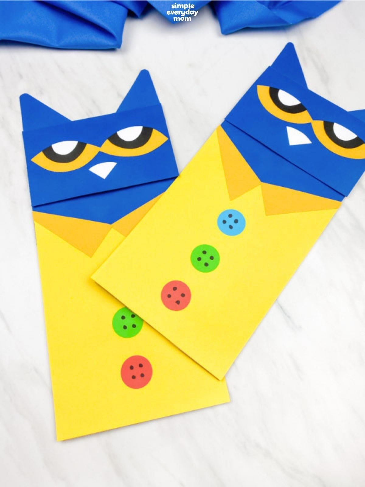 Two Pete the Cat paper crafts arranged diagonally on a white surface, each with a blue head, yellow shirt, orange collar, and colorful buttons.