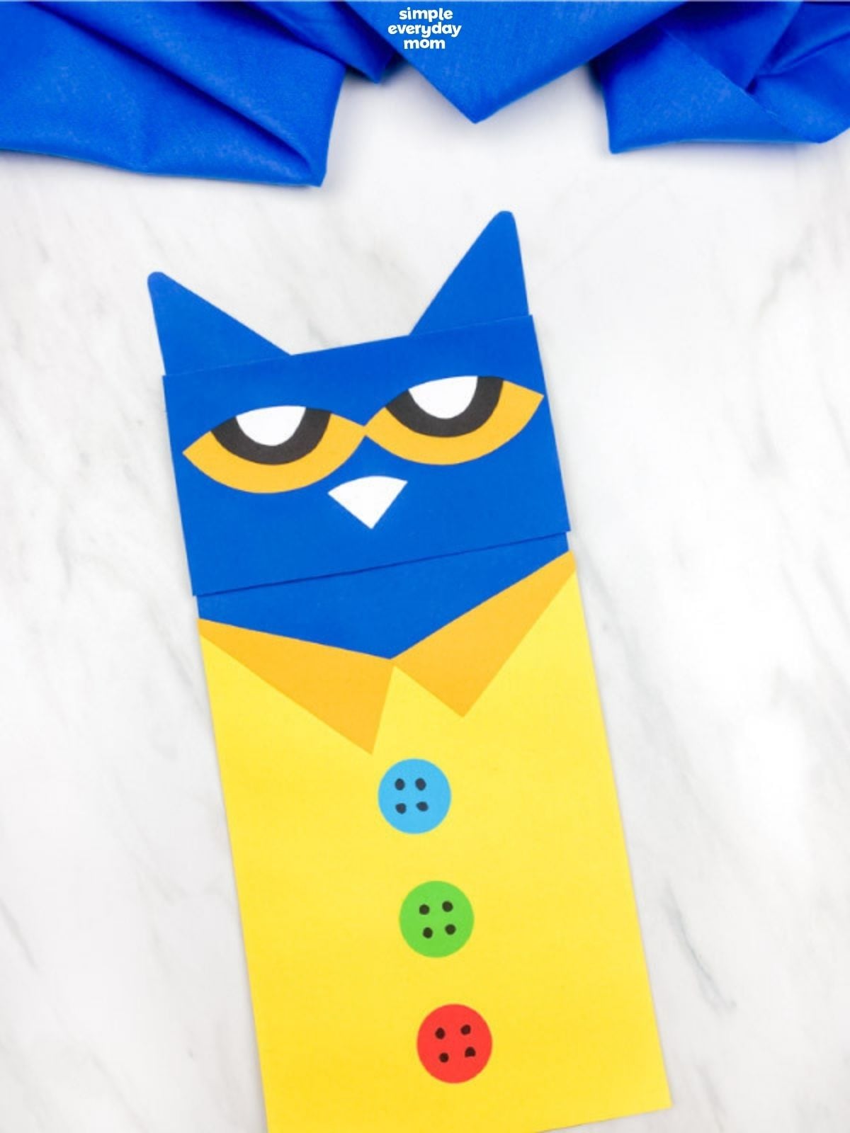 Single Pete the Cat paper craft lying on a white surface, with a blue head, yellow shirt, orange collar, and three colorful buttons.