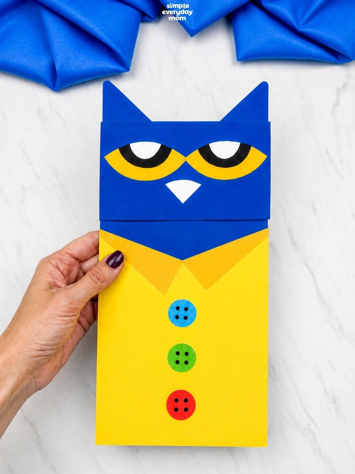 Hand holding a Pete the Cat paper bag puppet craft with a blue head, yellow shirt, orange collar, and three colorful buttons on a white surface.