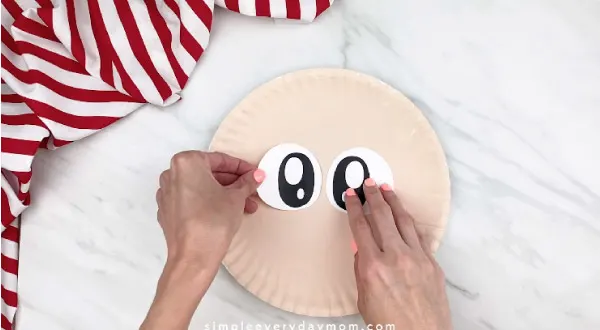hands gluing the eyes onto peach paper plate 