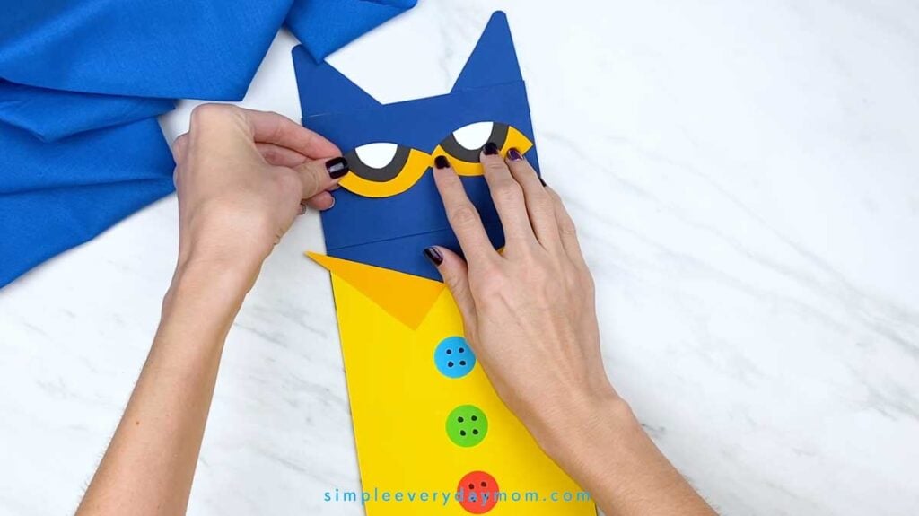 Hands position the completed yellow, black, and white eyes onto the blue face of the Pete the Cat puppet.