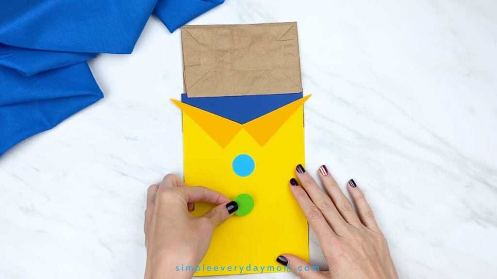Hands add colorful blue, green, and red paper buttons down the center of the yellow shirt.