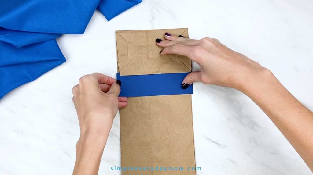 Hands attach a dark blue paper strip across the upper section of a brown paper bag to begin the Pete the Cat puppet