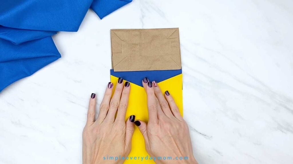 Hands press a yellow paper shirt piece onto the lower portion of the brown paper bag beneath the blue strip.